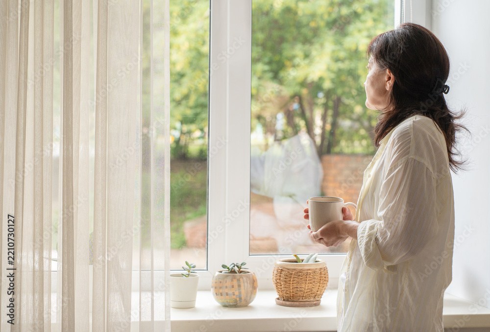 Fototapeta premium Modern adult woman in simple loungewear stands by window with cup of coffee, enjoying quiet moment of solitude. A slow morning ritual, an authentic moment in life of real person