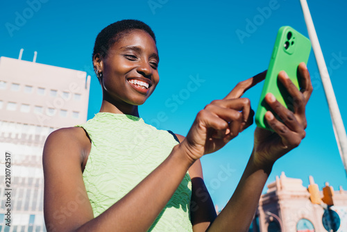 Joyful black woman using smartphone screen for text messaging or online chatting outdoors. Happy African American female traveler scrolling social network feed on bright urban background.