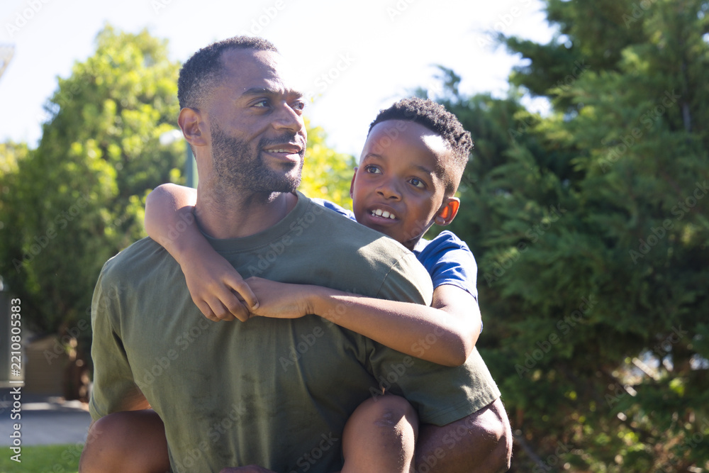 Fototapeta premium Father carrying youth son piggyback in yard wearing olive-green and blue tees smiling toward right