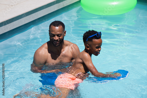 African American father and son practicing swimming in home pool with blue kickboard in trunks