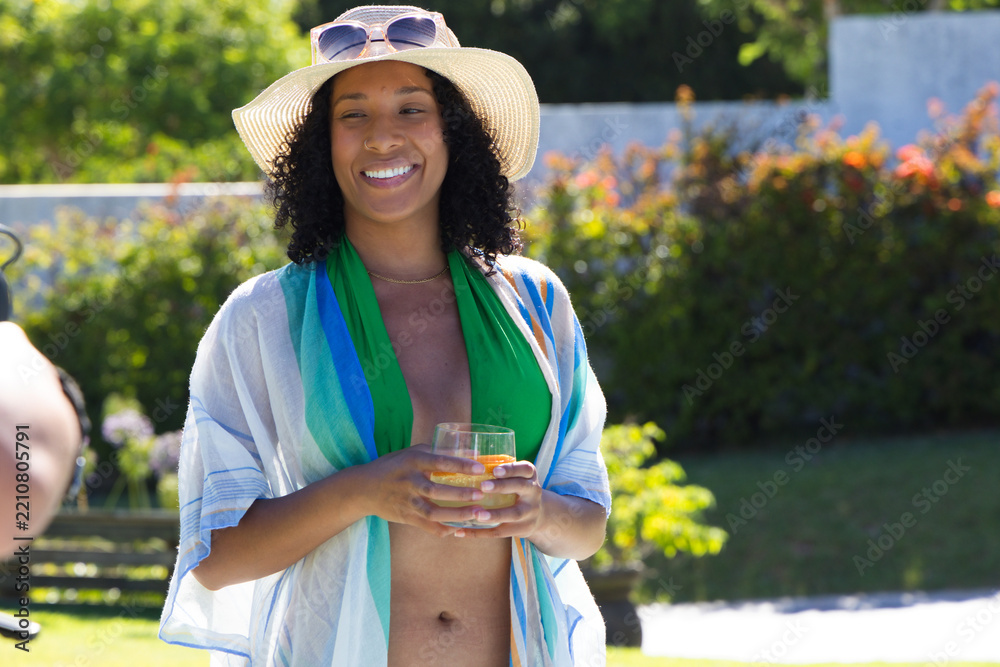 Obraz premium African American woman standing poolside in green swimwear straw hat holding orange cocktail glass