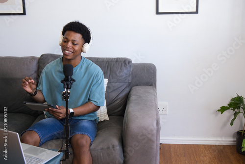 Adult African American man sitting on sofa in home studio recording with microphone, holding tablet