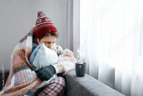 man freezing inside a cold home suffers from an energy crisis today. High heating costs, weak heating, and winter frost leaves him drinking a hot cup of tea to seek extra warmth.