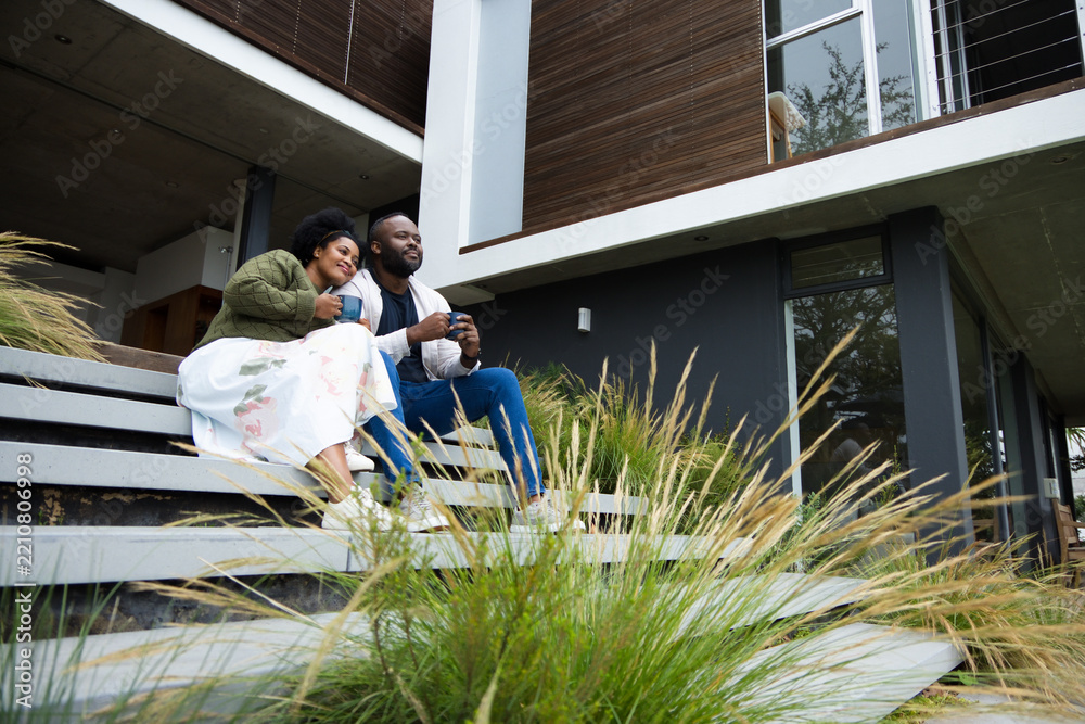 Obraz premium Couple sitting on wide horizontal steps outside modern two-story house, holding ceramic mugs