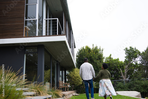 African American couple walking on lawn toward modern glass home, passing stone steps, copy space