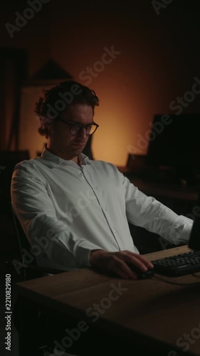 Suffering from pain young businessman working on computer late at night in dark office feels tired and stressed, experiences neck pain and burnout from overwork and long hours at desk, slow motion.