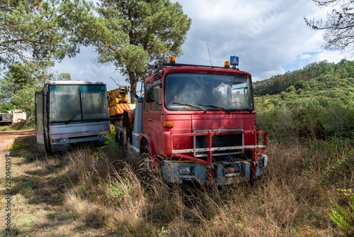 Weathered red heavy truck parked beside an abandoned passenger bus among tall dry grass and vegetation. Aging commercial vehicles represent obsolete transport, fleet retirement, industrial salvage, ve