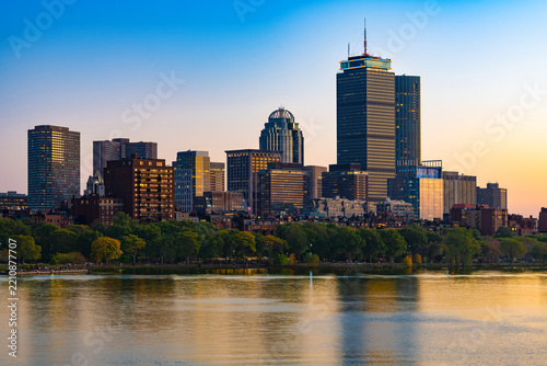 The urban skyline of the Back Bay neighborhood in Boston seen across the Charles River at sunset featuring tall architectural towers, colorful evening reflections, and tree lined riverfront banks.