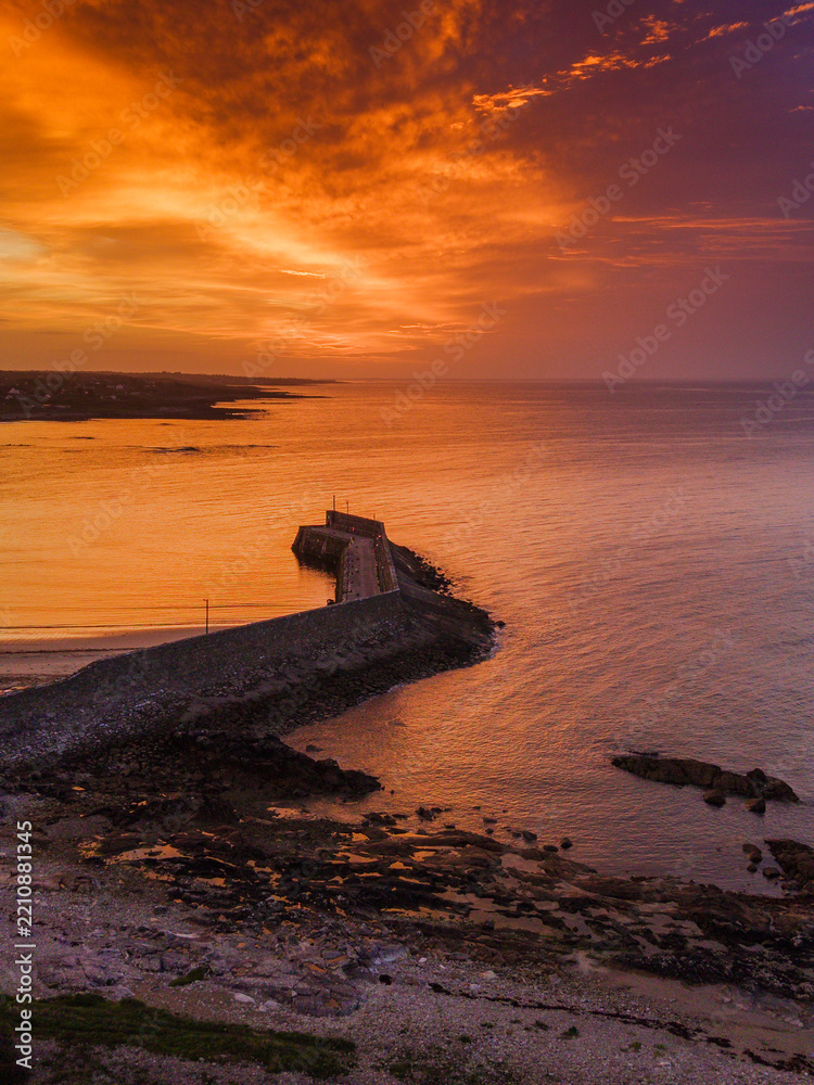 Fototapeta premium Aerial view of coastal pier at golden sunset. Warm light reflects on water and stone breakwater. Calm sea meets land with sandy beach nearby. Spiddal, Ireland, Galway bay. Travel and tourism.