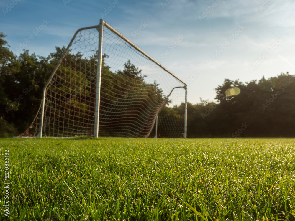 Naklejka premium White soccer goal stands alone on green grass. Trees line the background under blue sky with wispy clouds. Scene peaceful and quiet, no people present. Ideal for sports, recreation