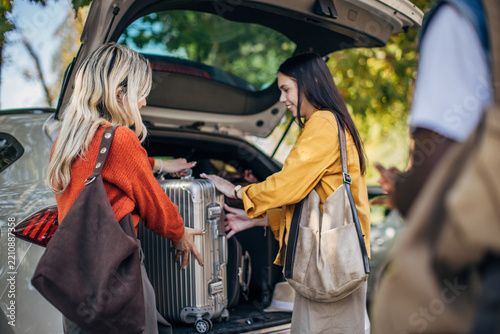 Under a sunny sky, two friends carefully place a large suitcase into the trunk of a car, preparing for their journey. The atmosphere is lively and filled with excitement as they organize their belongi