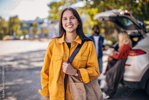 A young woman smiles at the camera while standing next to a car. Friends are getting ready for a road trip, showcasing a sense of adventure and camaraderie. Bright sunlight highlights the vibrant colo