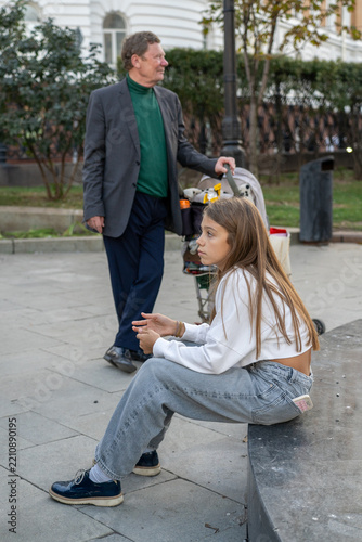 Young girl in casual clothing sits on stone ledge while older man stands behind her, holding onto stroller in outdoor urban setting.