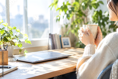 Young woman holding steaming coffee mug while working on laptop at home