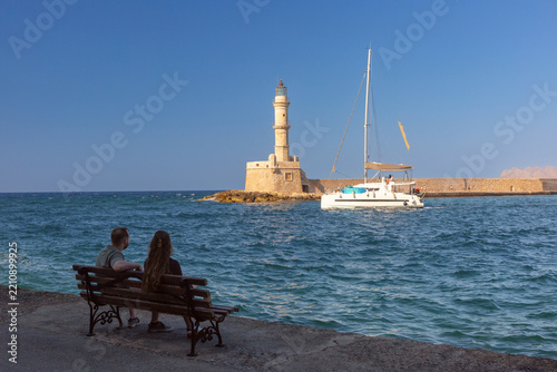 Couple sitting on a waterfront bench overlooking a sailboat and the historic lighthouse at the Old Venetian Harbor in Chania, Crete, Greece.