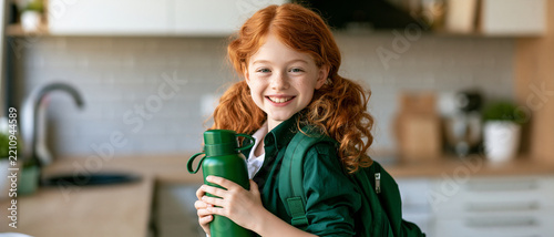 A happy girl with red hair and pigtails, holding a green water bottle and wearing a green backpack, smiling at the camera