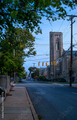 Historic stone church with a tall Gothic tower at a street intersection in Frederick, Maryland, featuring green trees and a blue morning sky.