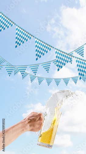 A person holds a large glass beer mug, spilling and splashing beer against a background of bright blue sky with white clouds and traditional Bavarian checkered pennants. Bavarian Oktoberfest Festival