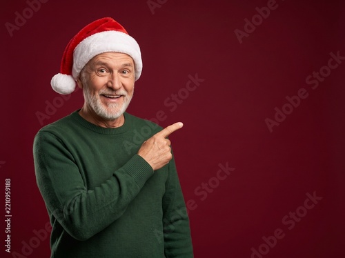 Excited senior man in Santa hat pointing finger at copy space while smiling on red background