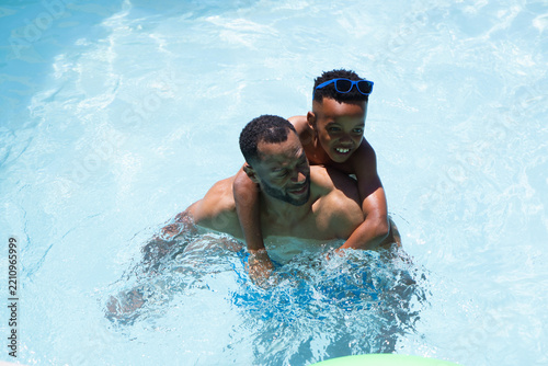 African American father and son splashing in sunlit pool with blue sunglasses, trunks, green float