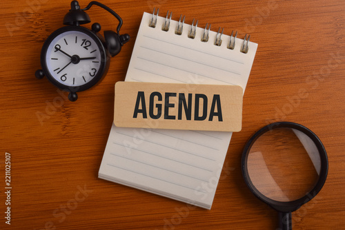 Agenda planning concept featuring a wooden agenda sign, notebook, alarm clock, and magnifying glass on a wooden desk, representing scheduling, organization, and time management.