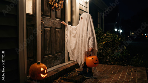 Child dressed as ghost holding pumpkin bucket knocking on door with festive background of Halloween night and glowing jack o lantern