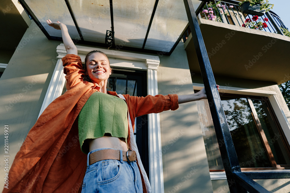 Obraz premium Happy young woman stretching arms with joy on the porch of a house at sunrise. Wearing an orange coat and green top. Concept of freedom, morning energy, and new day.