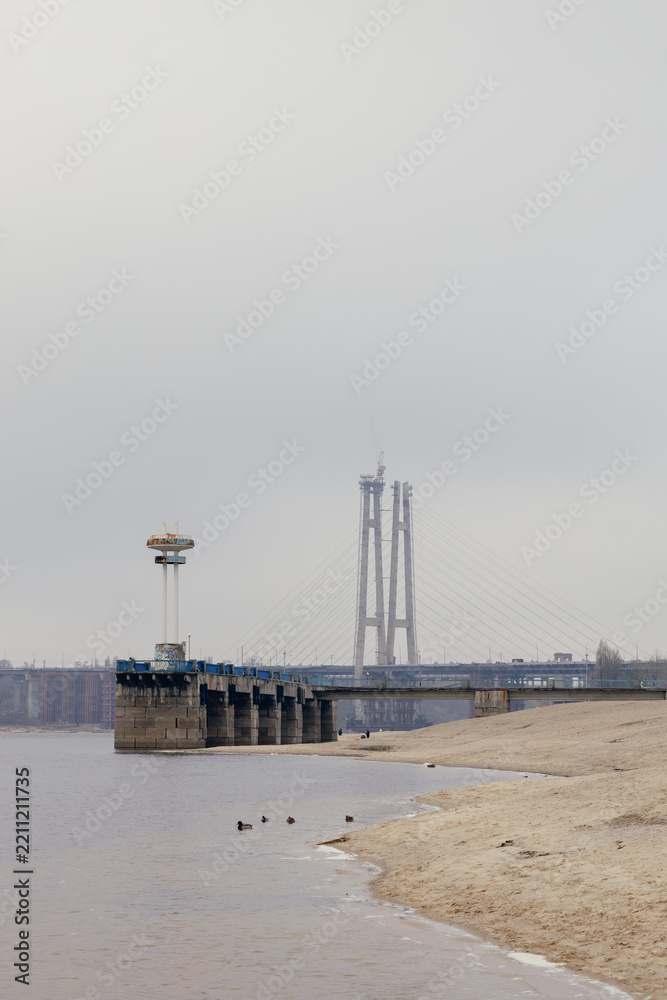 Fototapeta premium Gloomy riverfront with an old pier and a bridge under construction