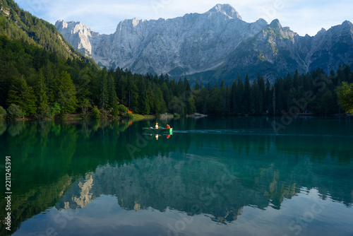 People rowing a boat on a serene mountain lake surrounded by dense forest and towering rocky peaks reflecting in calm water
