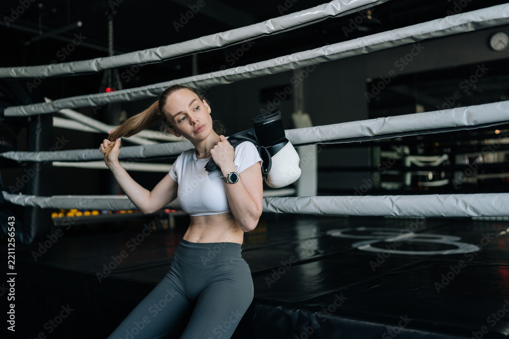 Obraz premium Female athlete holding boxing gloves and adjusting hair, taking break inside boxing ring after challenging workout, embodying dedication and strength in fitness journey.