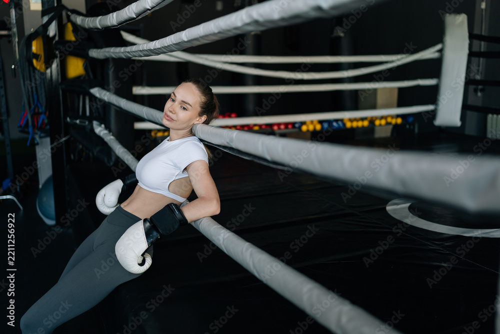 Obraz premium Woman athlete wearing boxing gloves leaning on ropes of boxing ring, taking break from training in gym environment. Concept of on fitness journey and strength, healthy sports life.