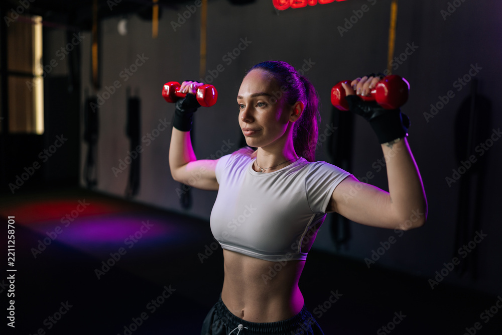 Obraz premium Focused woman with muscular body training with dumbbells in fitness club. Low key lighting with purple and red accents creates motivated atmosphere for strength training and bodybuilding.