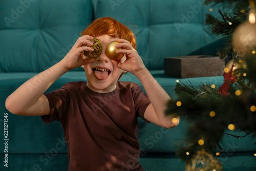 A cheerful boy holds Christmas baubles over his eyes at home