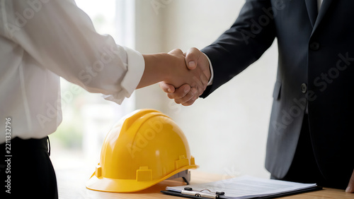 Business professionals shaking hands over a desk with a yellow hard hat and clipboard