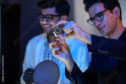 A researcher presents the die that came off the wafer while a colleague leans in to look with him inside a dimly lit research and development laboratory late in the working day