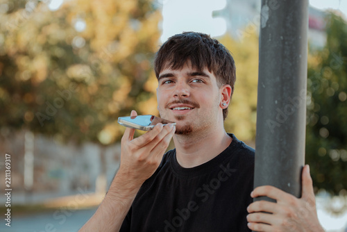 Young Caucasian man smiling while using voice assistant on smartphone outdoors, standing by a lamppost in an autumn park setting