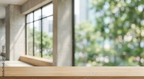 Empty light wooden table top in foreground with blurred modern interior background featuring large black frame windows and green trees outside, bright daylight