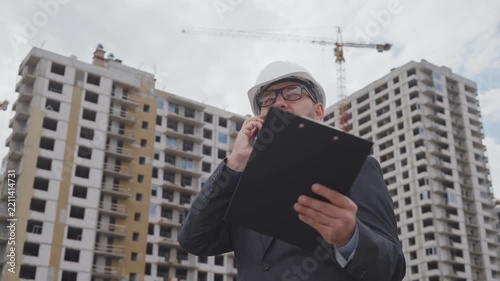 Successful male architect in suit talking on smartphone while holding clipboard at building site. Business engineer supervising project