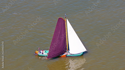 Aerial view of a traditional Dutch sailing boat with a maroon mainsail and white jib gliding across the water with people on deck in Heeg, Friesland, Netherlands.