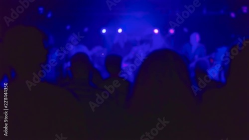 Blurred crowd at live concert in nightclub under blue and purple stage lights. Audience silhouettes near illuminated music stage in dark club. Defocused indoor performance with colorful spotlights