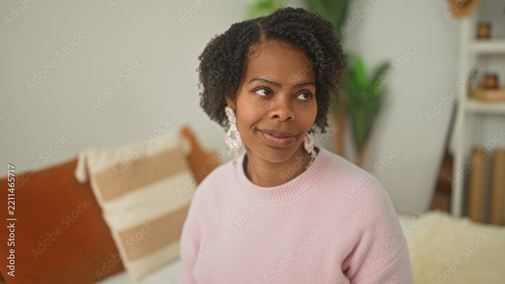 Fototapeta premium Young woman on bed wearing pink sweater and statement earrings, eyes closed with pursed lips beside pillows and plant in building; calm mindfulness.