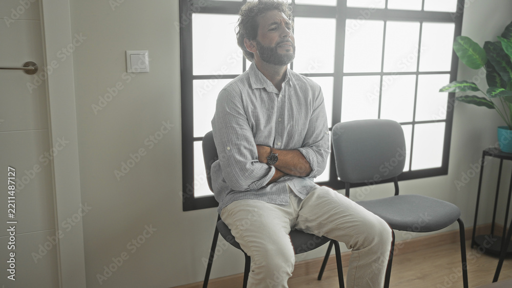 Fototapeta premium Man sitting on chair with arms crossed and hands clasped over stomach in building waiting area near window and plant; anxiety waiting.
