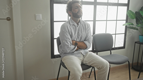 Man sitting on chair with arms crossed and hands clasped over stomach in building waiting area near window and plant; anxiety waiting.