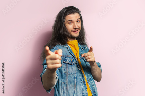 Young happy man with long hair pointing index finger on camera. Isolated on pastel pink background.