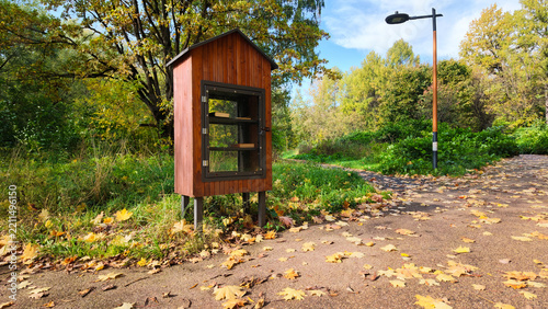 A small wooden library stands beside a walking path in a park. The library has a glass front and is surrounded by trees and fallen leaves. It is a sunny day in autumn.