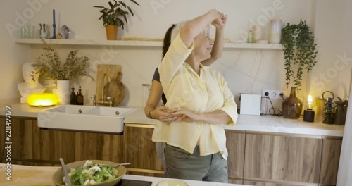 Joyful elderly mother and adult daughter dancing and laughing together while preparing a meal at home