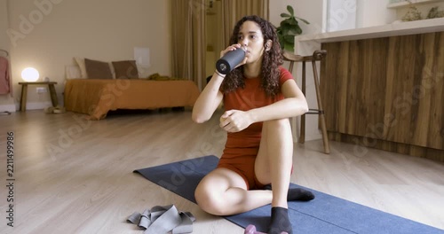 Young athletic woman drinking water from a bottle while sitting on a yoga mat after exercising at home