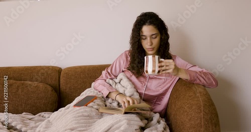 Relaxed young woman in pajamas reading a book and using her smartphone while sitting on the couch