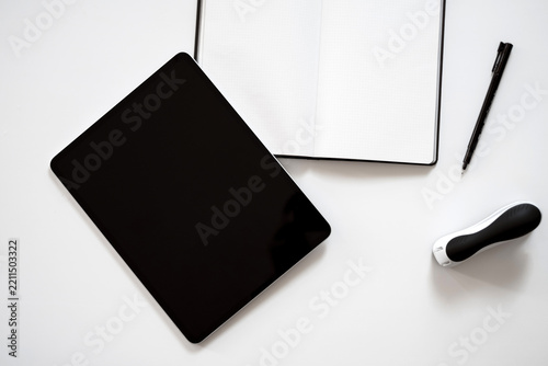 Minimalist workspace with tablet, notebook, and pen. White surface highlights clean, organized creative tools. Stylish black tablet sits beside open journal and pen