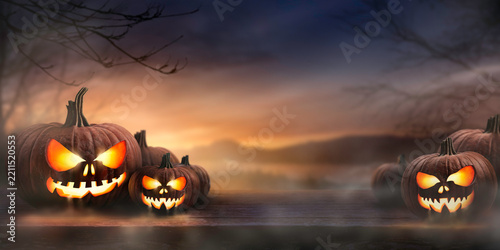 A spooky sunset with the haunted evil glowing eyes of Jack O' Lanterns on the left and right of a wooden bench with copy space centre on a scary halloween night